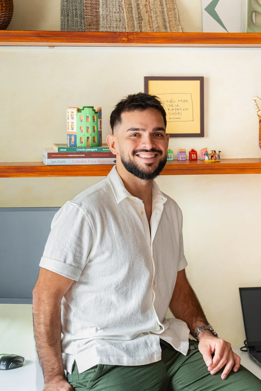 Giann Freire, arquiteto, sorrindo no escritório, de camisa branca, diante de uma estante com livros