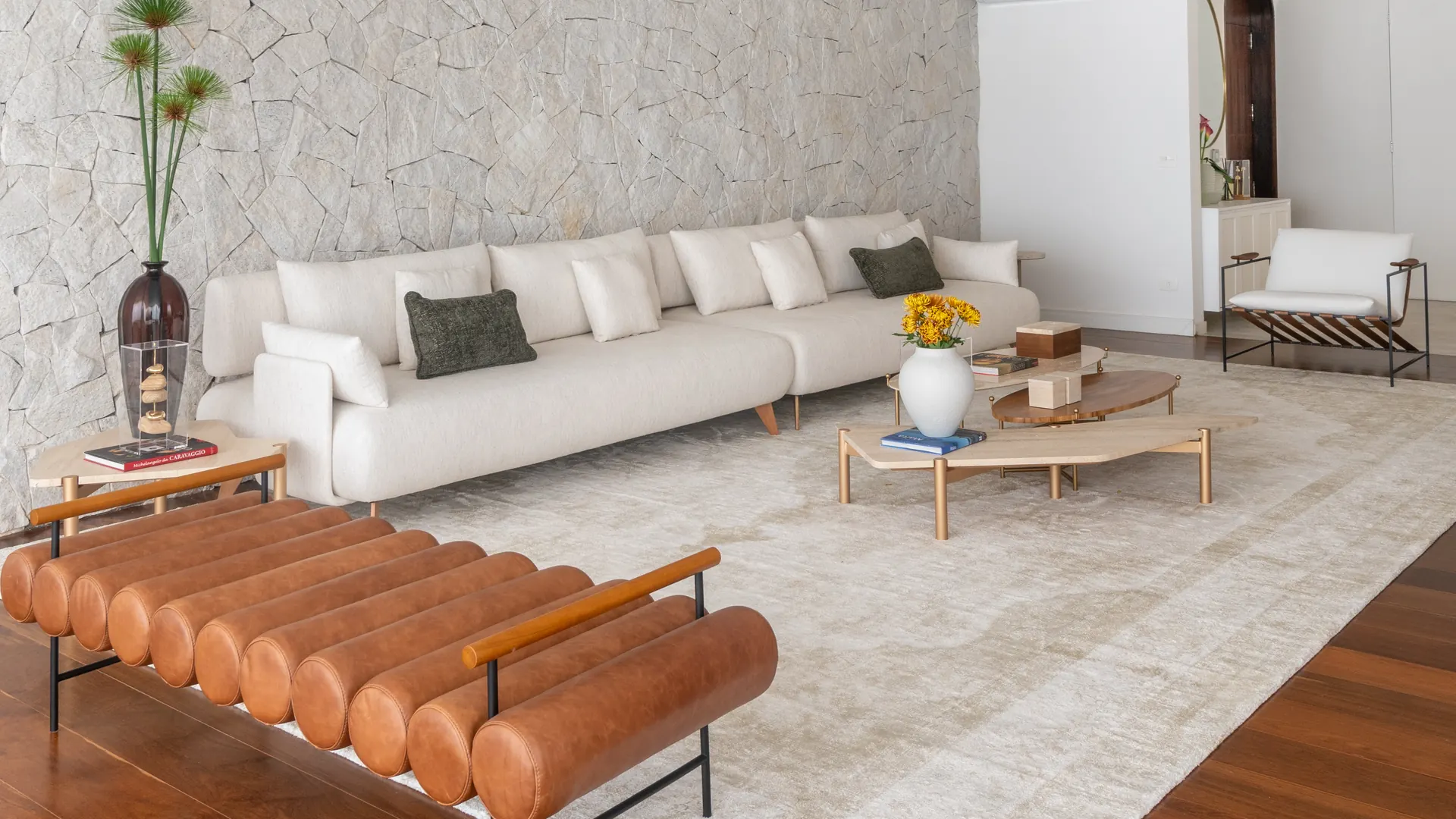 Sala com parede de pedra natural, sofá claro e banco de couro caramelo